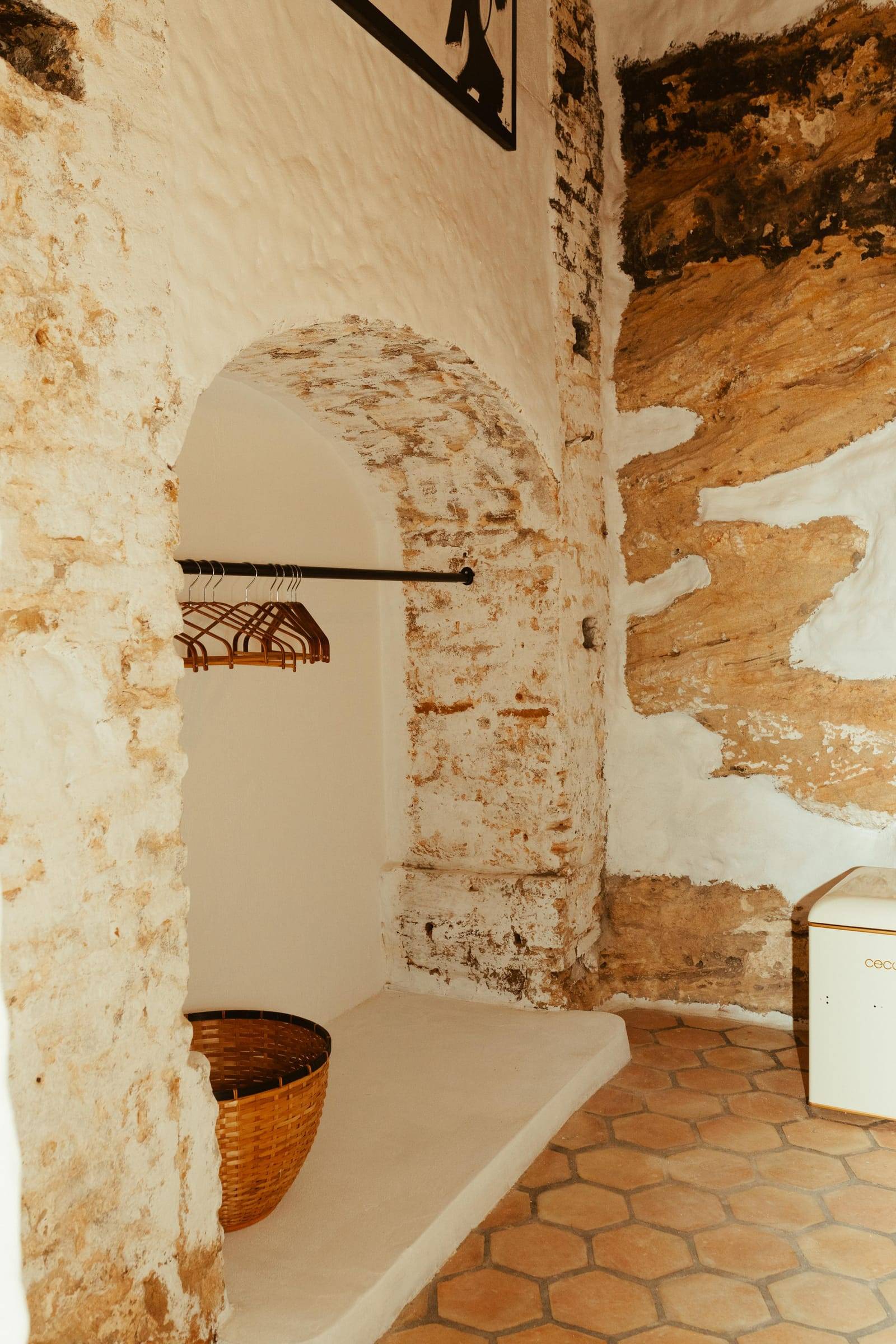 Hornacina encalada bajo un arco de ladrillo, con barra para la ropa, cesta de mimbre y suelo de barro