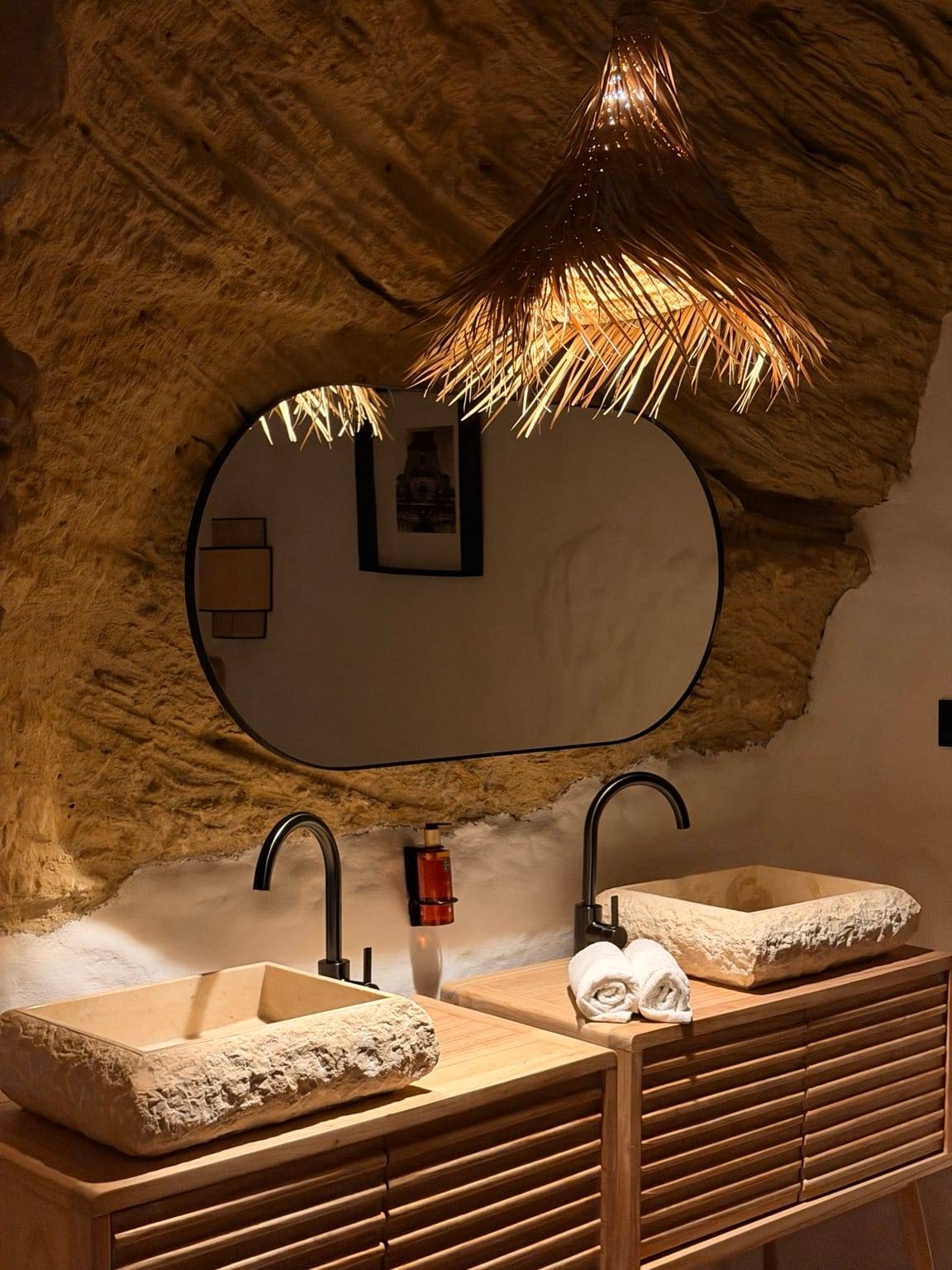 Baño excavado en la roca con dos lavabos de piedra sobre un mueble de madera, espejo ovalado y lámpara de fibra natural
