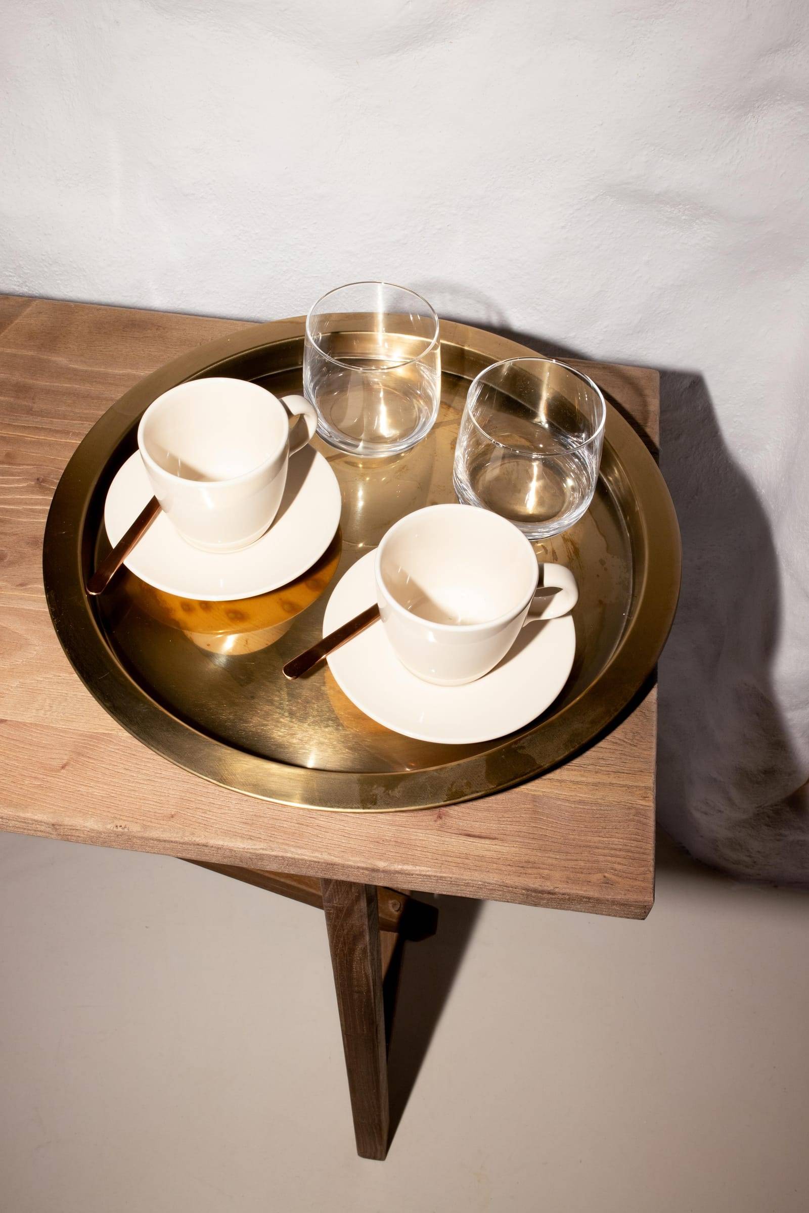 Dos tazas de café y dos vasos de agua en una bandeja dorada, sobre un banco de madera junto a una pared encalada