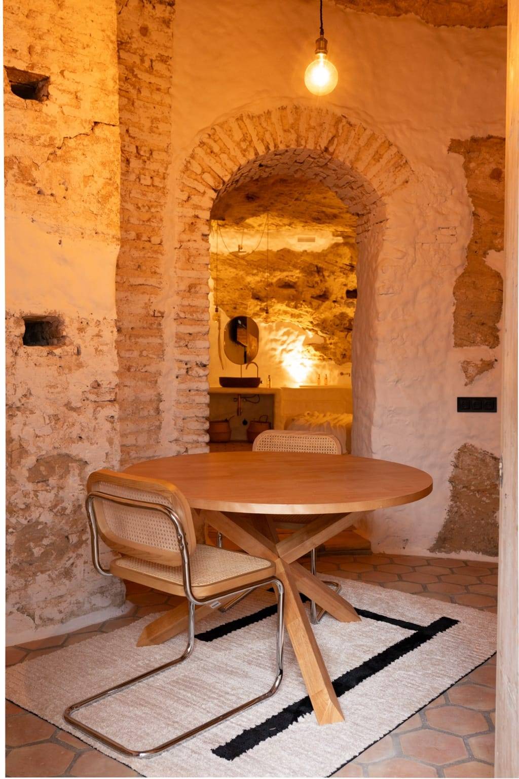 Mesa redonda de madera con sillas de rejilla ante un arco de ladrillo que se abre a una estancia iluminada