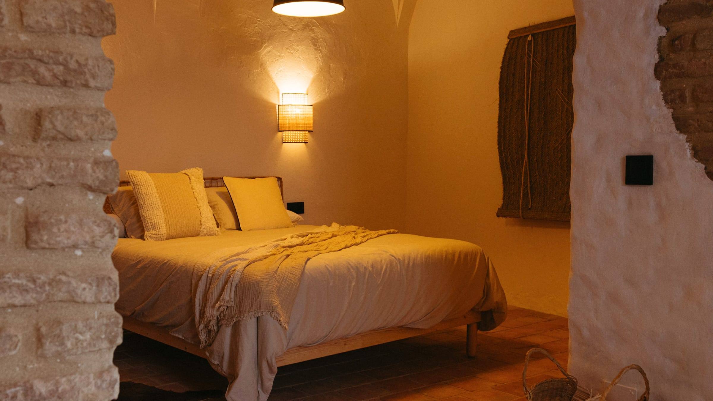 Dormitorio con luz cálida, pared de ladrillo visto y cama vestida en tonos claros