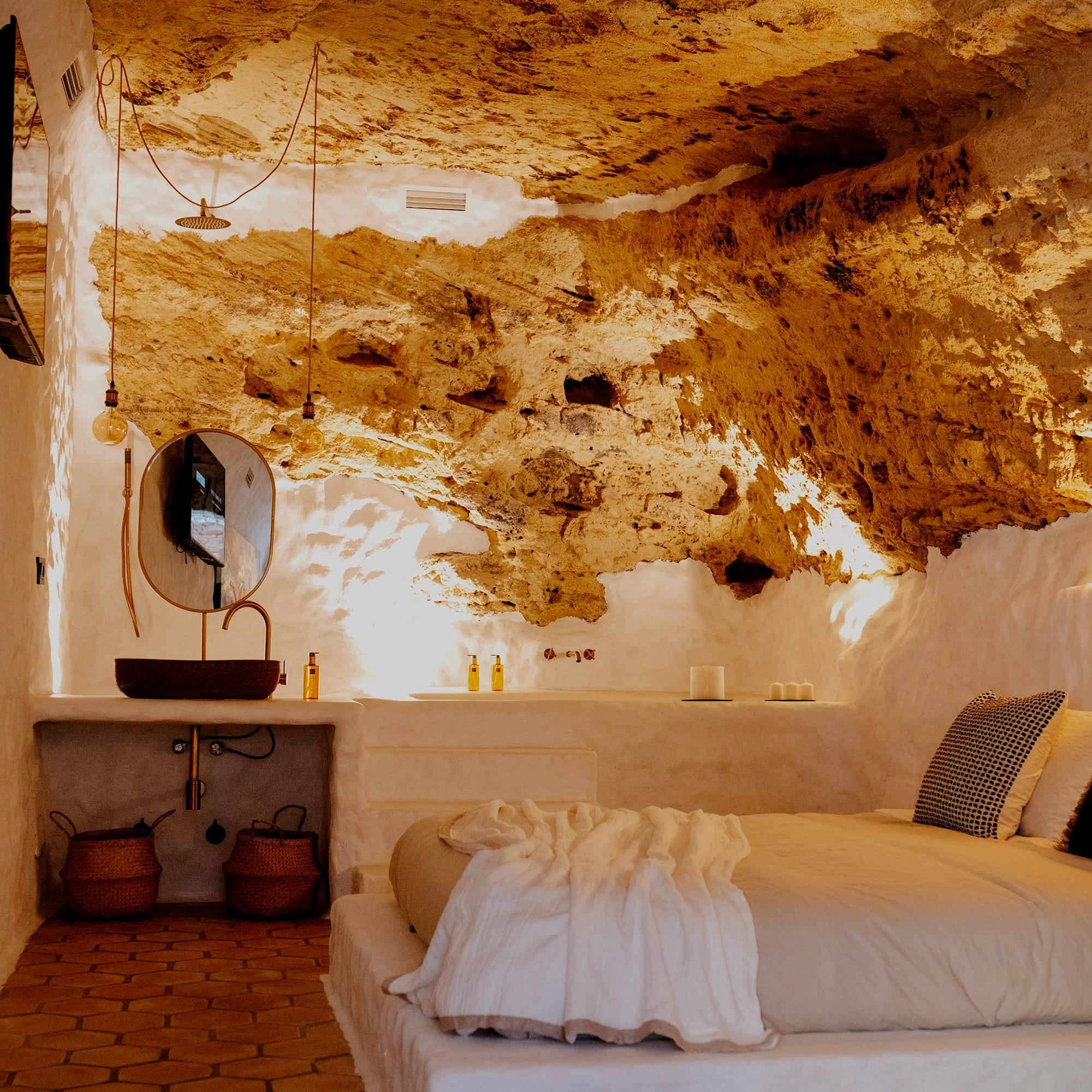 Dormitorio bajo un techo de roca natural, con cama blanca, lavabo de piedra y espejo ovalado