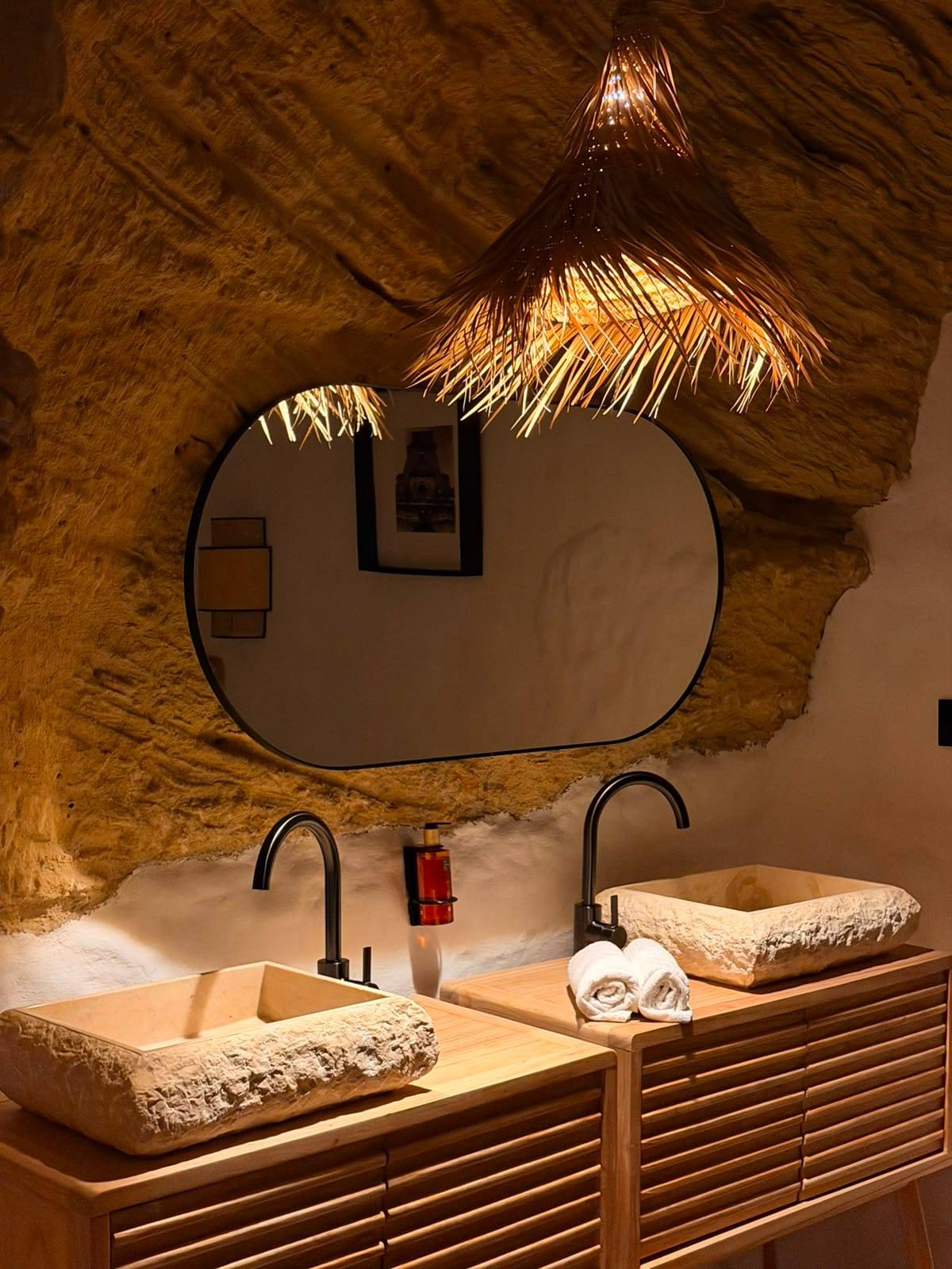 Dos lavabos de piedra sobre muebles de madera, con espejo ovalado y lámpara de fibra natural bajo el techo de roca