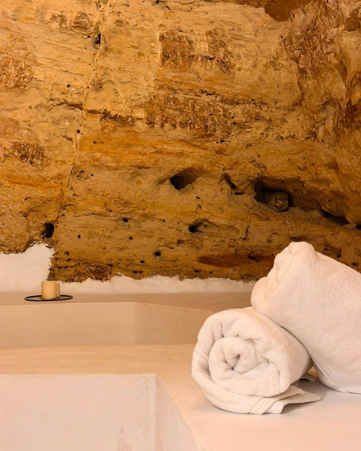 Toallas blancas enrolladas y una vela junto a la bañera excavada, bajo el techo de roca